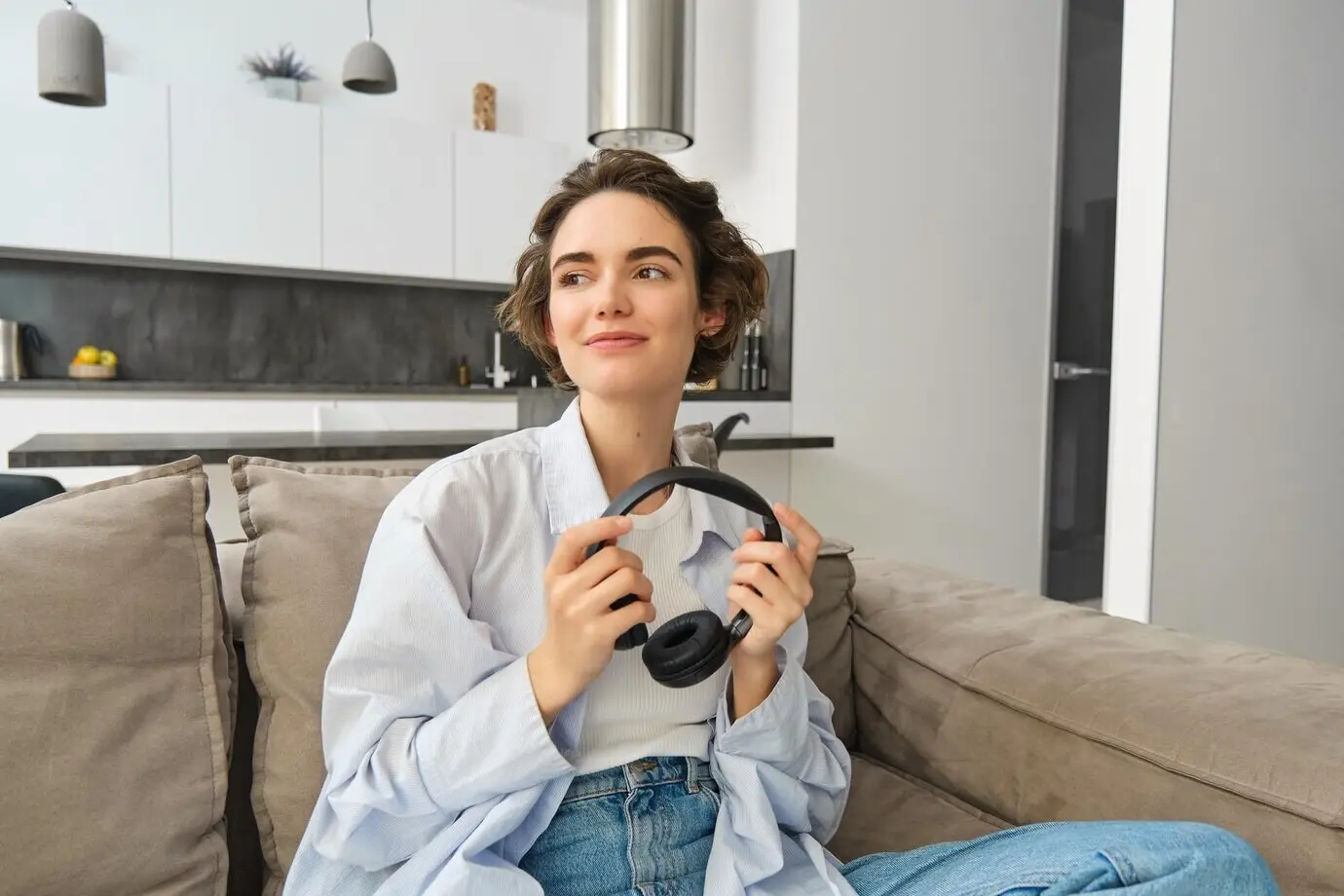 Porträt eines schönen, modernen Mädchens, das auf einem Sofa sitzt, Kopfhörer in den Händen hält, zur Seite blickt und zuhört.